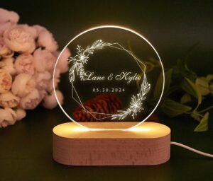 A clear acrylic LED lamp with a circular design, featuring an etched floral wreath and geometric border. Inside the wreath, the names "Lane & Kylie" are inscribed in a elegant script, along with the date "05.30.2024". The acrylic disc is illuminated by a warm light from its oval wooden base, creating a glowing effect. In the soft-focus background, there are pink flowers and green leaves, adding to the romantic and celebratory ambiance.