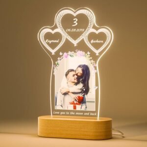 A beautifully illuminated, personalized acrylic lamp stands on a light wooden base, serving as a heartfelt commemorative gift. The acrylic panel is shaped with a large central arch framing a romantic photo of a couple embracing, with the man holding a gift. Above the photo, three interconnected hearts are etched: the top heart displays "3" and "06.28.2019", while the two smaller hearts flanking it contain the names "Raymond" and "Barbara" respectively, all glowing with soft light. The arch around the photo is adorned with delicate floral and butterfly etchings, and at the very bottom, the loving message "Love you to the moon and back" is inscribed. The lamp emits a warm glow, creating a tender and personalized ambiance.