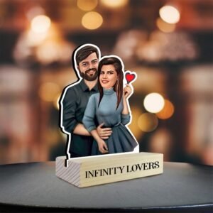 A custom caricature-style tabletop photo stand featuring a couple. The man is depicted with a beard, wearing a dark shirt, with his arms around the woman, who has dark hair and is wearing a blue top and skirt, holding a small red heart. The cutout of the couple is inserted into a wooden base that reads "INFINITY LOVERS." The item is placed on a dark, reflective surface, with a blurred background of warm, out-of-focus lights, creating a soft, romantic ambiance.