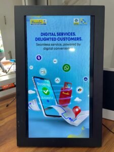 A vertically oriented digital display or light box showcasing an advertisement for "Bharatgas" digital services. The screen features a light blue background with text that reads "DIGITAL SERVICES. DELIGHTED CUSTOMERS. Seamless service, powered by digital convenience." Below the text, there's a graphic of a smartphone with "Booking Successful!" displayed on its screen, accompanied by a checkmark. A red LPG cylinder, branded "Bharatgas," is prominently featured, along with a map icon marked with a red location pin, suggesting delivery. Various digital payment and communication app icons, such as UPI, Paytm, and WhatsApp, are also scattered across the screen, indicating different service options. The top of the screen shows the Bharatgas logo and possibly another company logo. The display is framed in black and stands on a wooden surface, with a blurred background showing glimpses of a room.