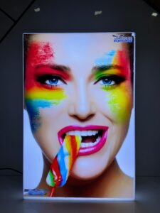 A vibrant, illuminated light box features a close-up image of a woman's face with striking blue eyes and bright pink lipstick. Her face is adorned with streaks of rainbow-colored paint, and she is playfully biting into a colorful lollipop, with some of the melted candy dripping from her mouth. In the top right corner of the light box, a logo with the word "FORTUNE" is visible. The light box itself is rectangular and emits a bright white glow, contrasting with the dark background.